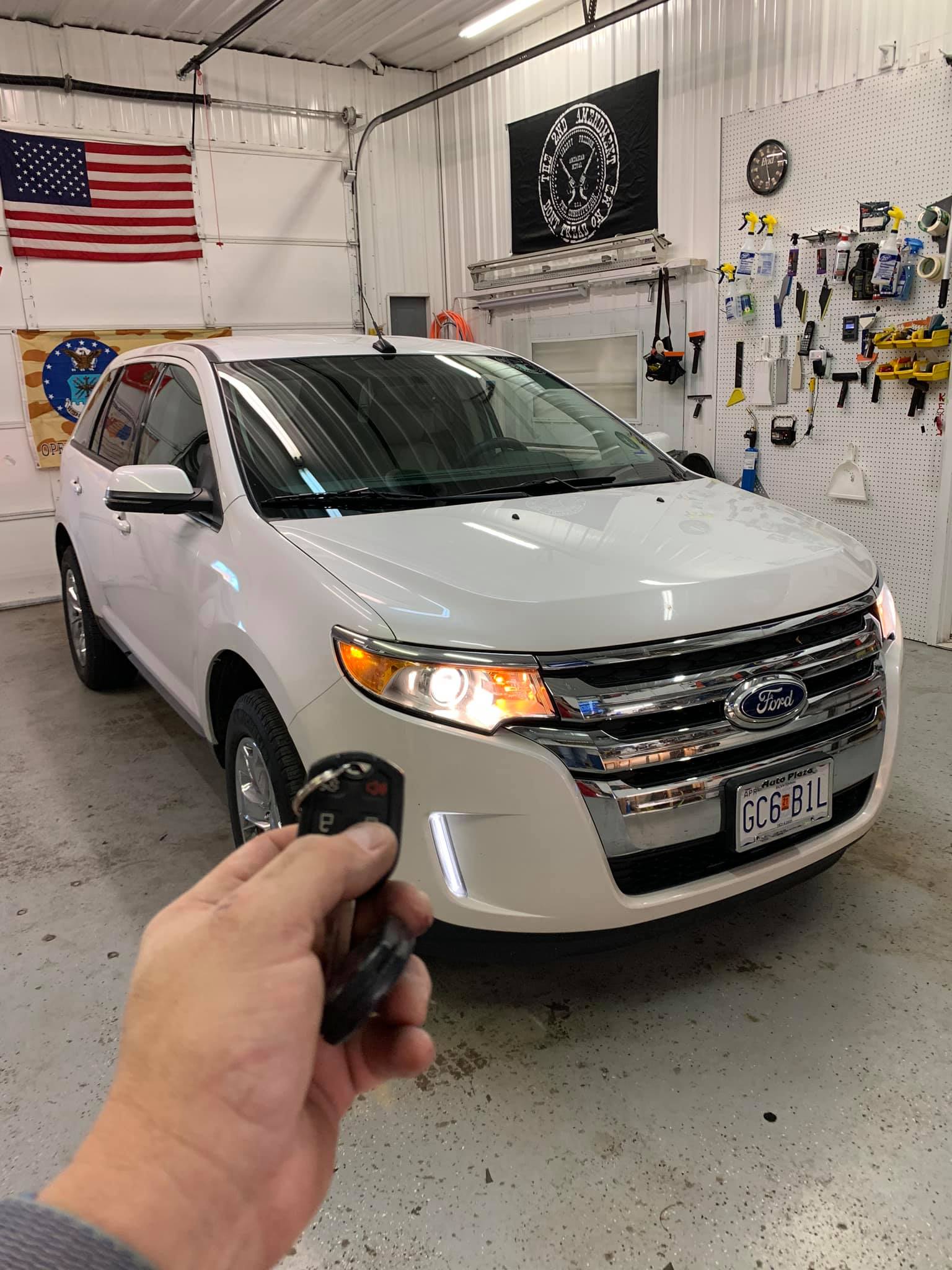 Hand holding car key fob toward a white SUV in a garage, with tools and an American flag on the wall