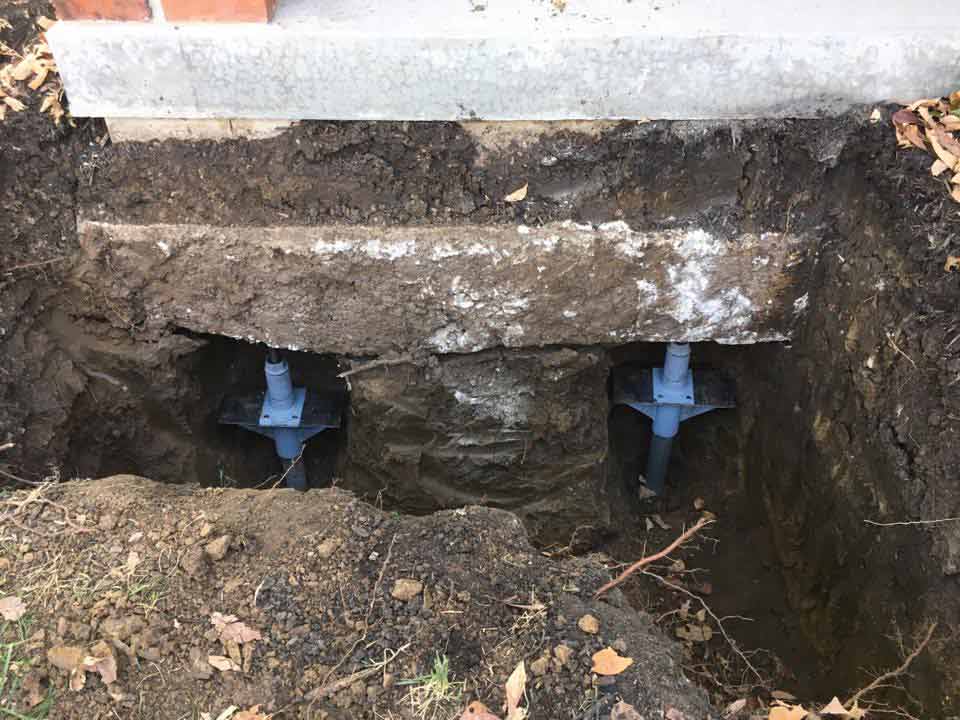 Two helical piers supporting a concrete foundation. Soil surrounds the piers in a dug-out hole.