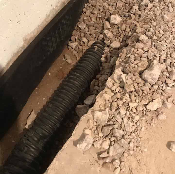 Black corrugated drain pipe in a trench next to a foundation wall with gravel and a black membrane.