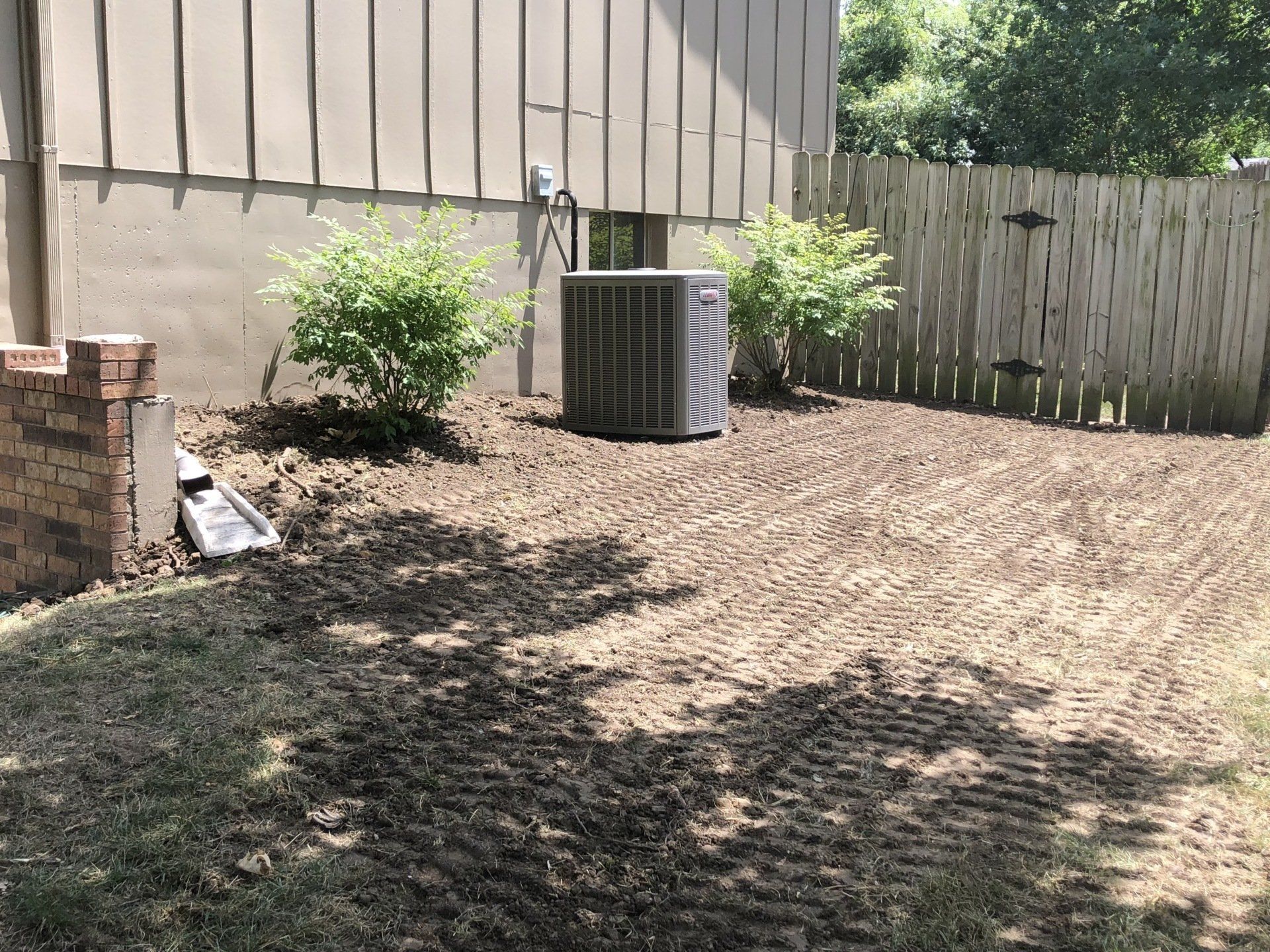 A yard with an air conditioner unit, two bushes, and a fence. The ground is tilled.