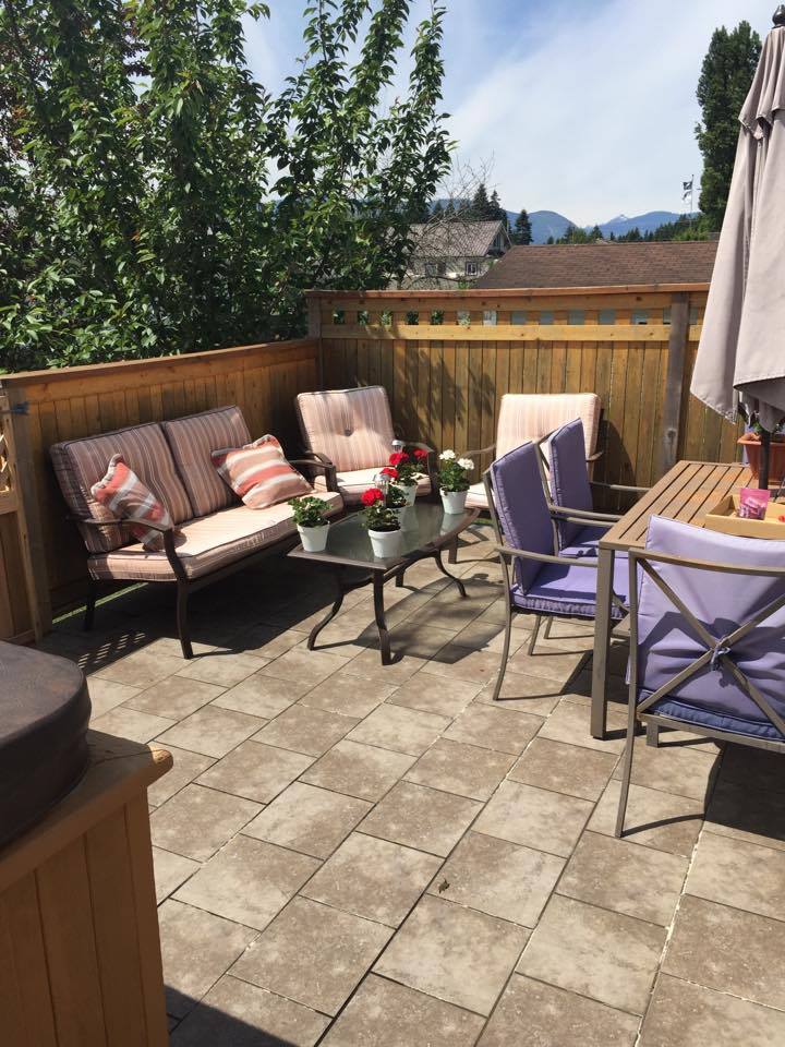 Patio with outdoor furniture: couch, chairs, table, and potted flowers. Wooden fence, tile floor, trees, and sky.