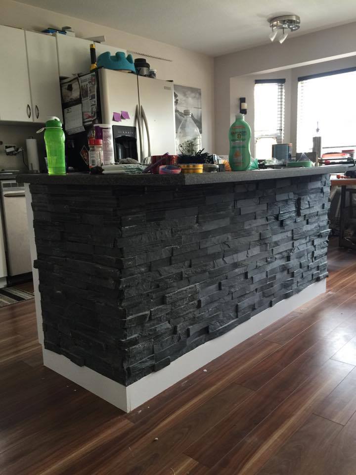 Kitchen island with black stacked stone facade, white base, wood flooring, and cluttered countertop.