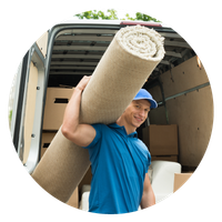A person in a blue shirt carries a rolled-up carpet toward a moving truck, smiling.