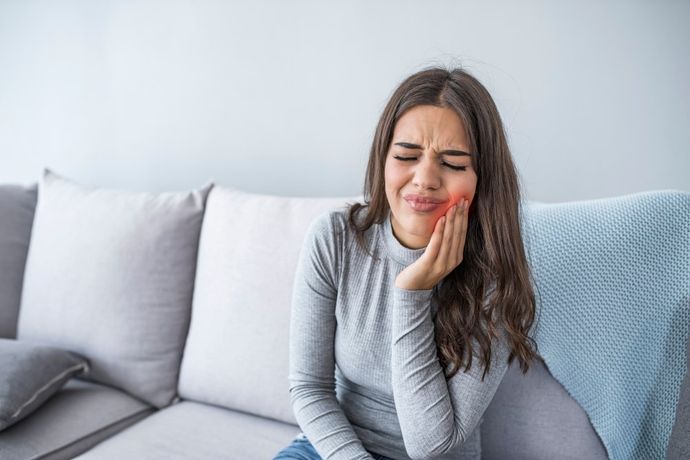 Femme sur un canapé avec une main sur sa mâchoire, montrant une expression de douleur ; son visage est rouge.