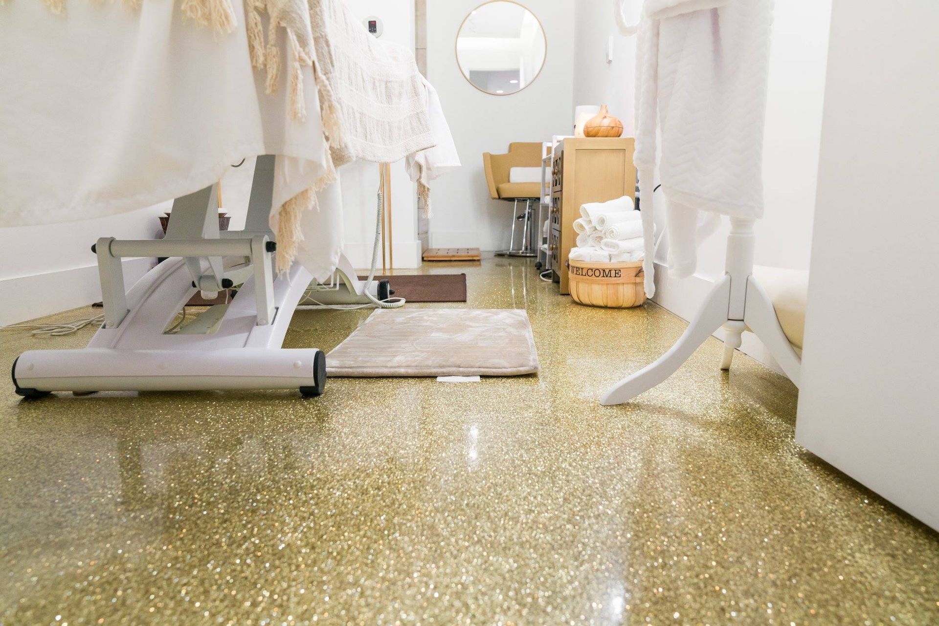 Bright spa room with white treatment tables, round mirror, shelves, and gold-speckled floor