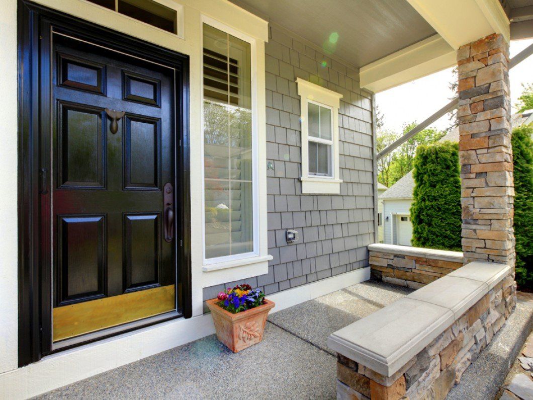 A black front door with a gold kick plate, beside a vertical window and grey shingle siding on a porch with stone accents.