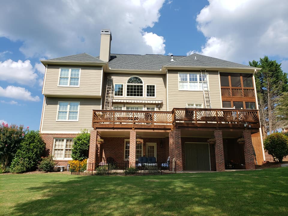 A two-story tan house with a brick lower level, featuring a wooden deck, screened porch, and a grassy backyard.
