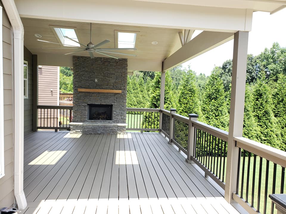 A spacious covered wooden deck features a stone fireplace, a ceiling fan, and black railings overlooking a treed yard.