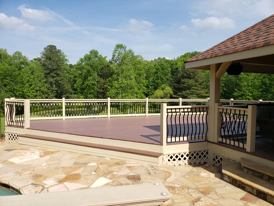 A spacious deck with a tan railing and dark metal balusters, overlooking a forested yard next to a stone patio.