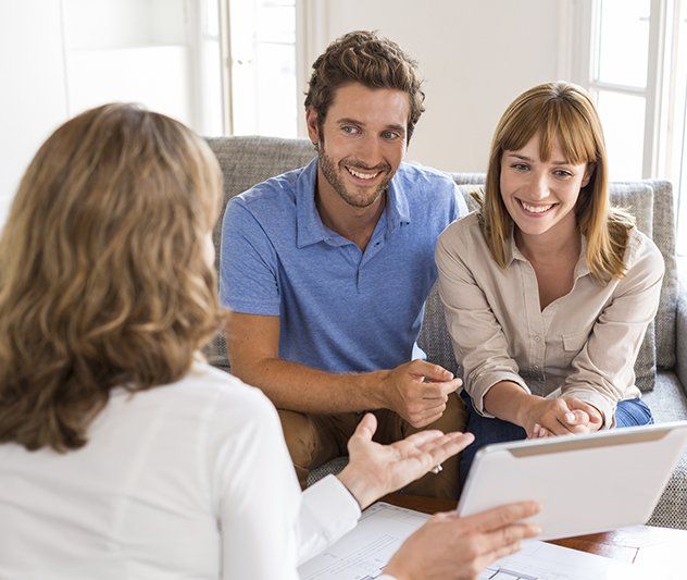 A man and woman are sitting on a couch talking to a woman while looking at a tablet.