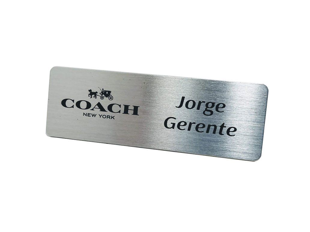 Placa de identificación plateada con las inscripciones “COACH” y “Jorge Gerente” sobre fondo blanco.