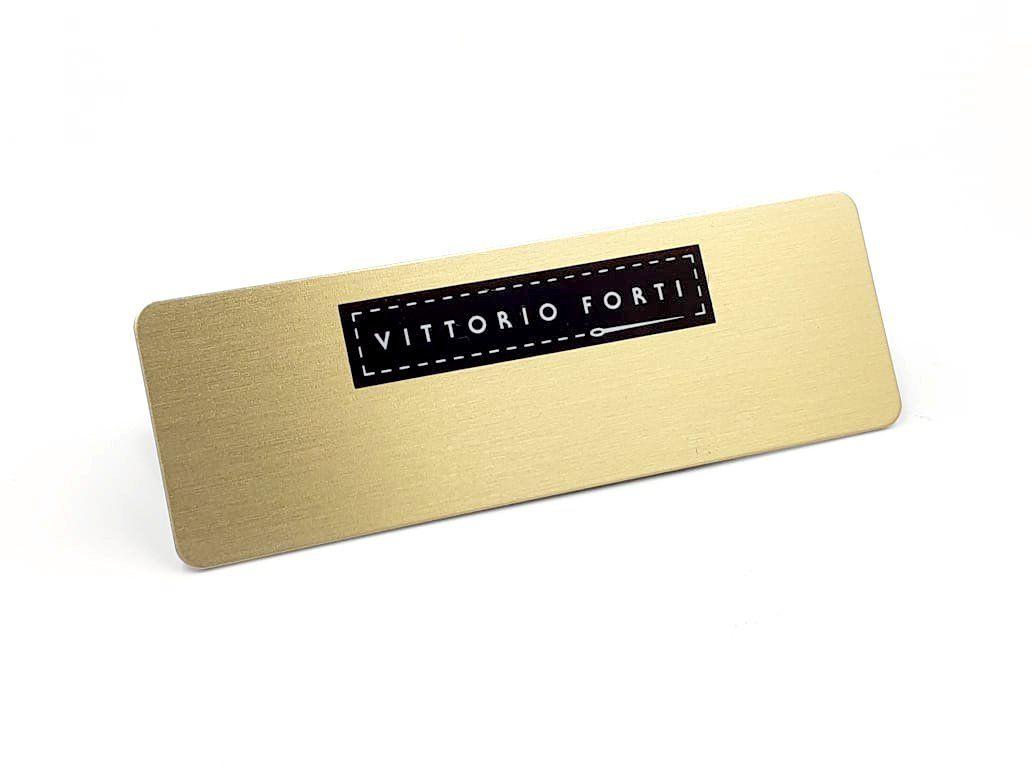 Placa rectangular de metal dorado con una etiqueta negra “Vittorio Fr…” centrada en ella.