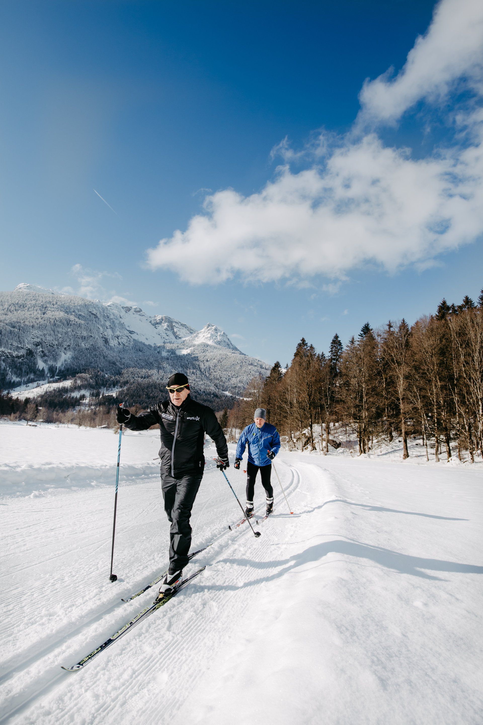 Zwei Skifahrer auf einer verschneiten Piste mit Bergen, Bäumen und blauem Himmel