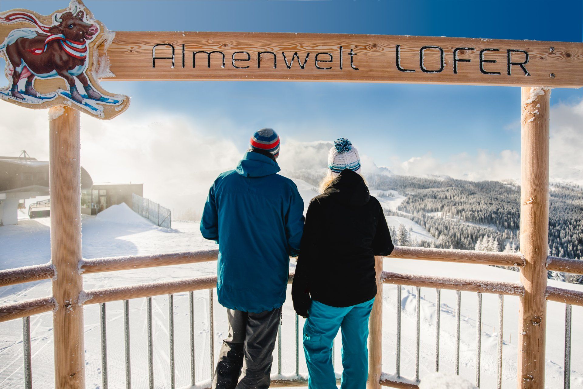 Zwei Personen in Winterjacken stehen auf einem verschneiten Aussichtspunkt unter einem Holzschild und blicken auf die Berge.