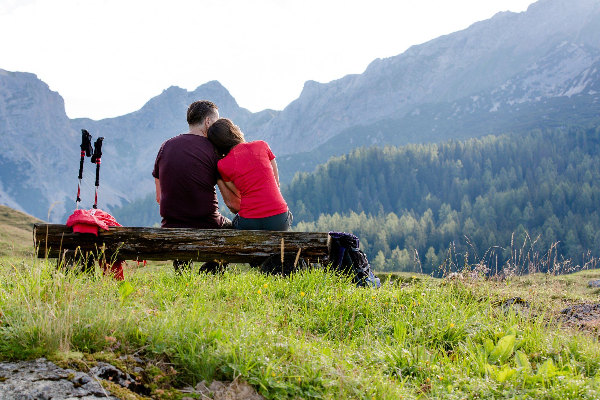 Zwei Personen sitzen auf einer Bank mit Blick auf ein grasbewachsenes Bergtal; neben ihnen steht ein roter Rucksack.