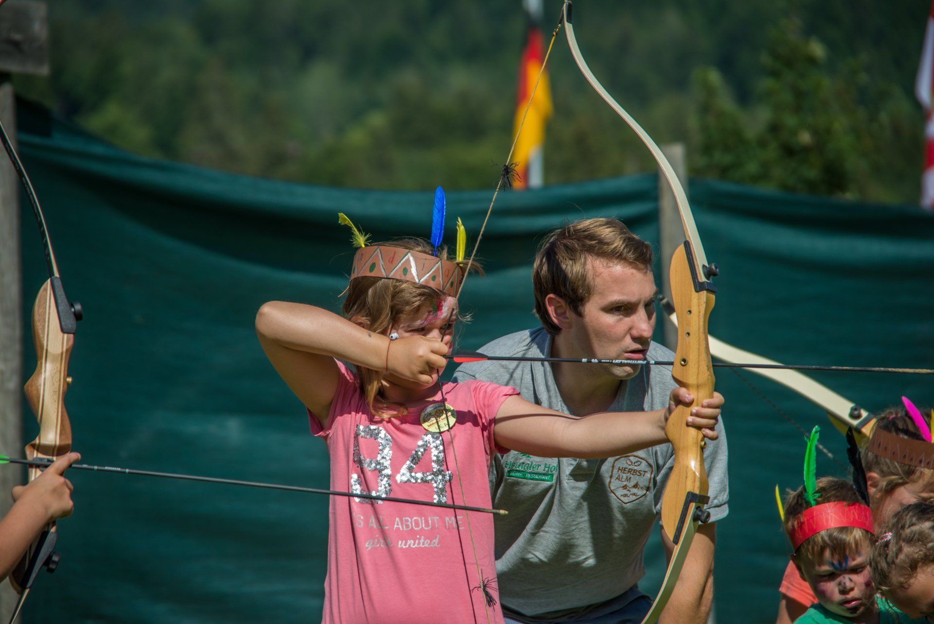 Bogenschützen zielen im Freien mit ihren Bögen; im Hintergrund ein Kind in Rosa und ein Erwachsener vor grünem Hintergrund.