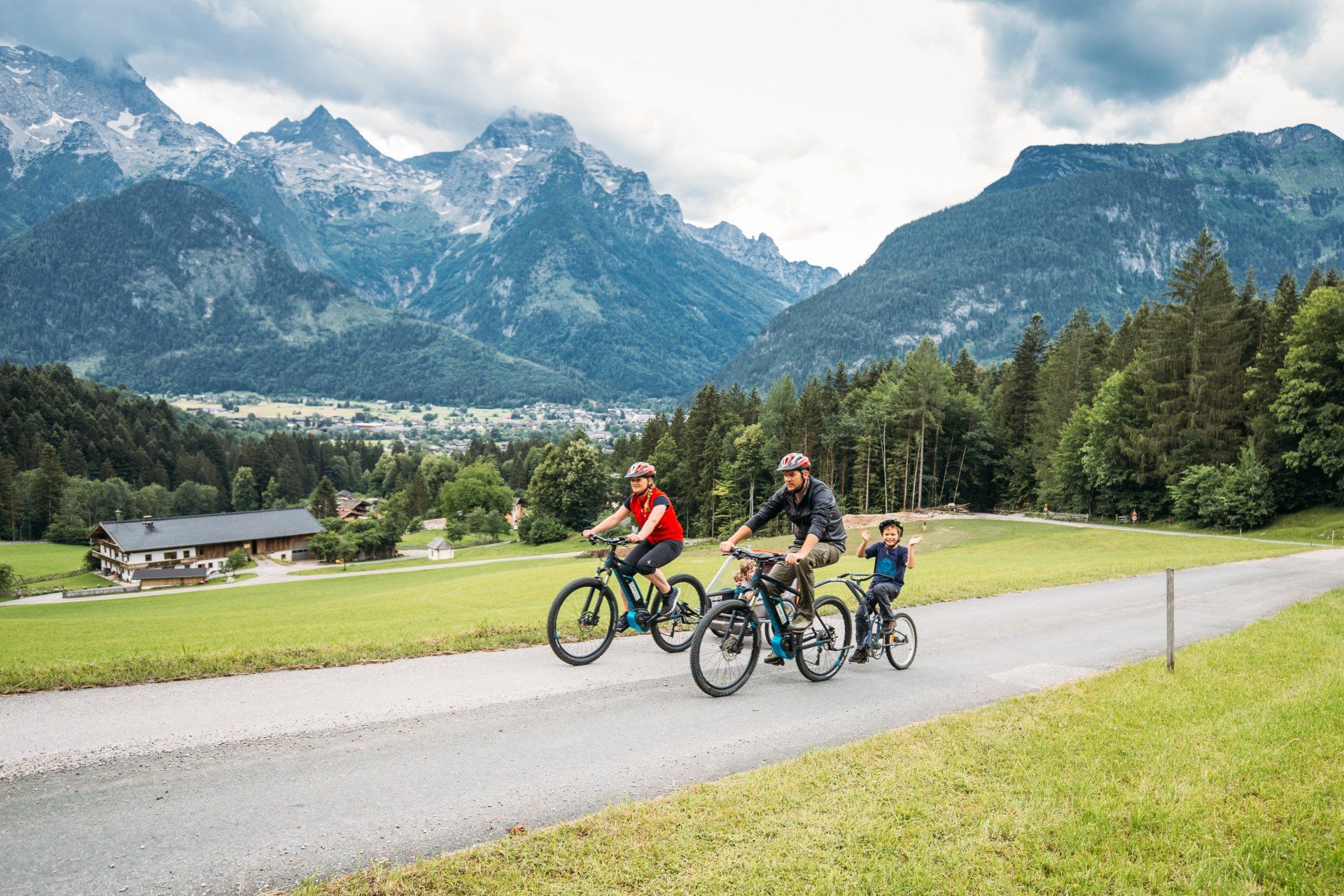 Radfahrer fahren auf einer Bergstraße mit schneebedeckten Gipfeln und einem bewaldeten Tal im Hintergrund