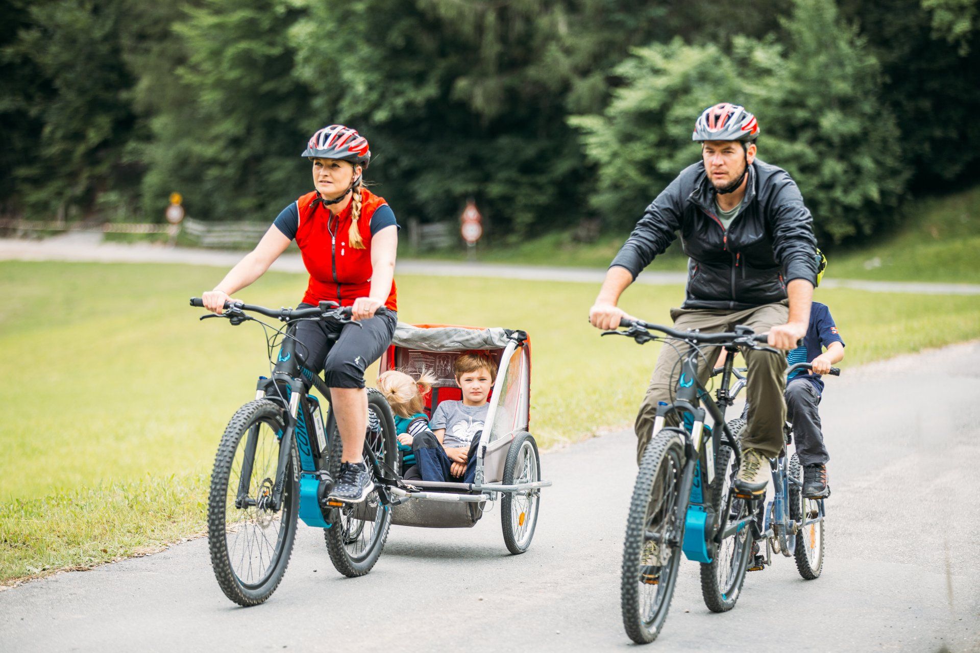 Zwei Radfahrer ziehen einen Kinderanhänger auf einem gepflasterten Weg durch einen grasbewachsenen Park.