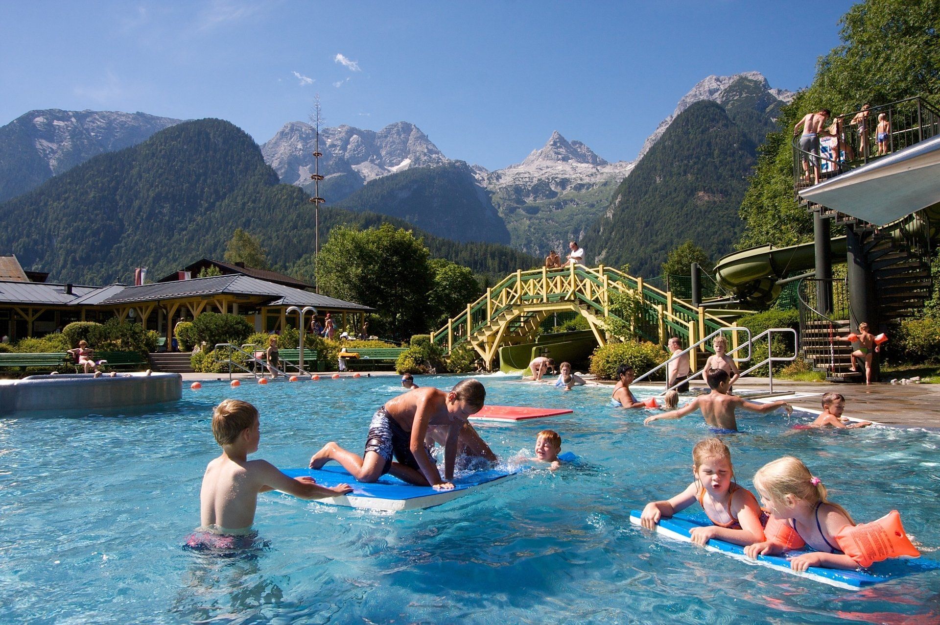 Kinder spielen in einem Hotelpool vor der Kulisse von Bergen und sonnigem Himmel.