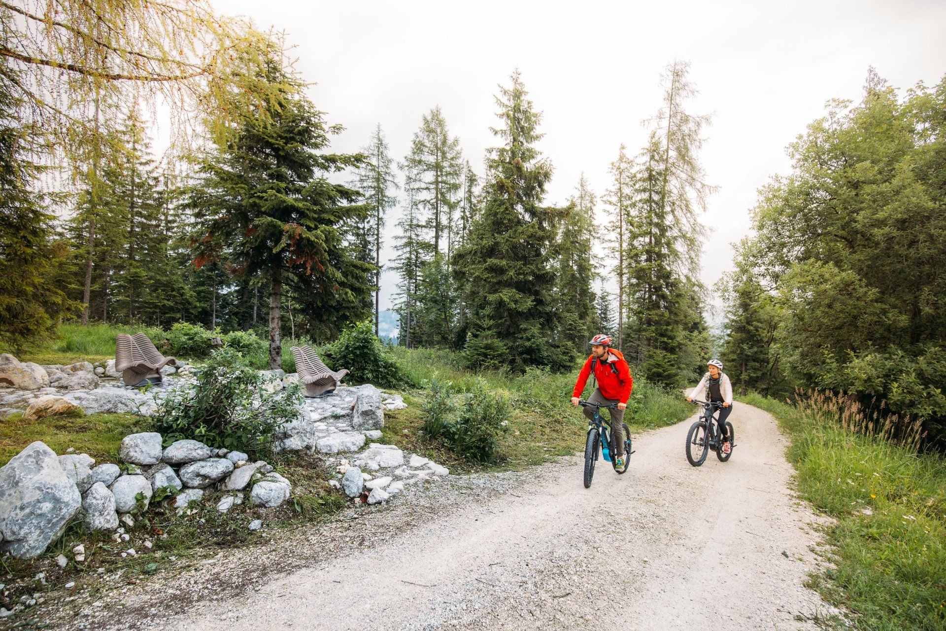 Zwei Radfahrer fahren auf einem Schotterweg im Wald neben Bäumen und Felsen.