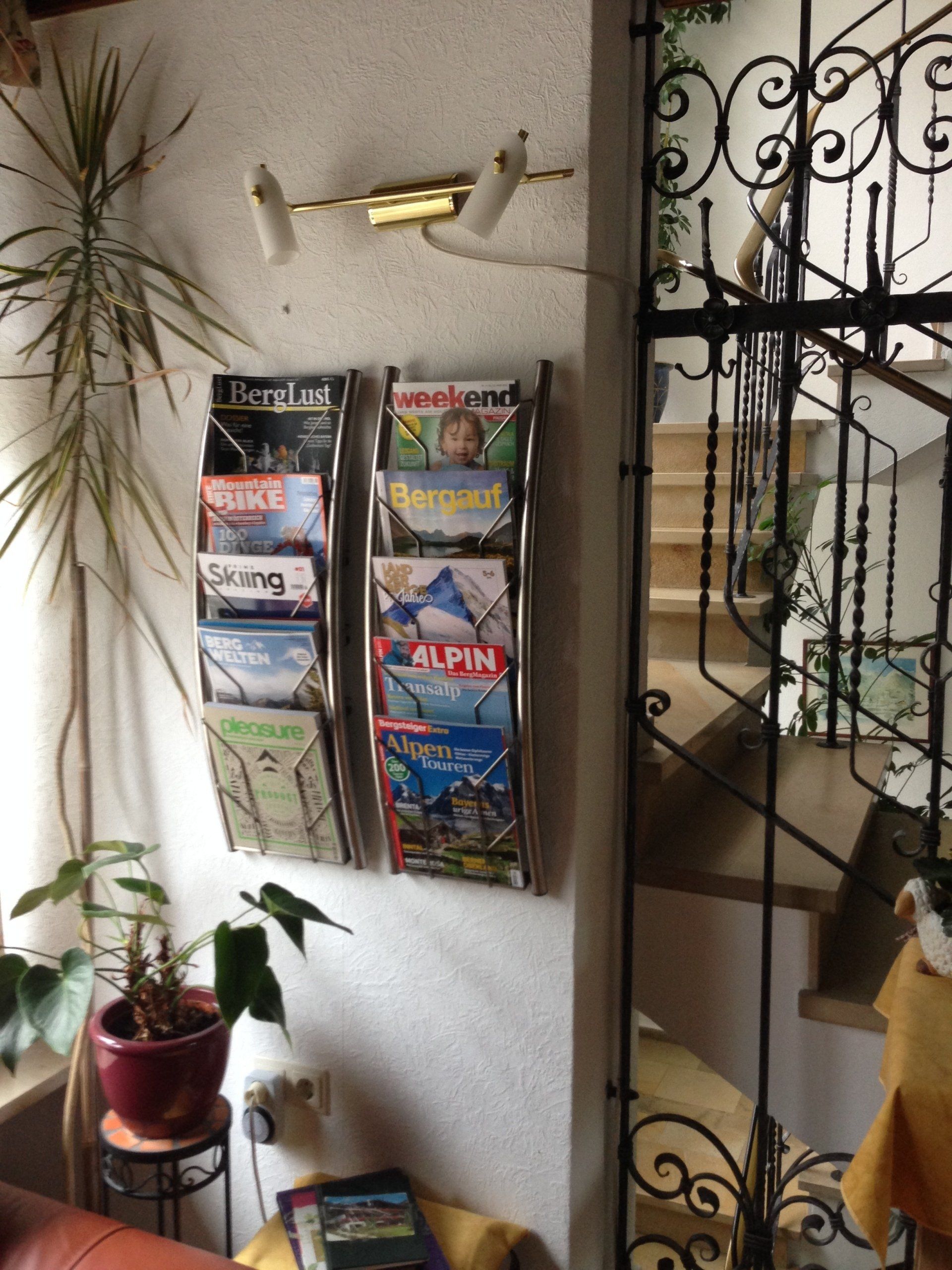Zeitschriftenständer an der Wand neben einer Topfpflanze und einem verzierten schwarzen Metalltürrahmen im Innenbereich.