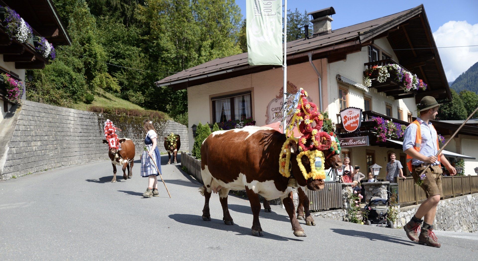 Eine mit Blumen geschmückte Kuh spaziert durch eine Dorfstraße, vorbei an Gebäuden im Chalet-Stil und Fußgängern.