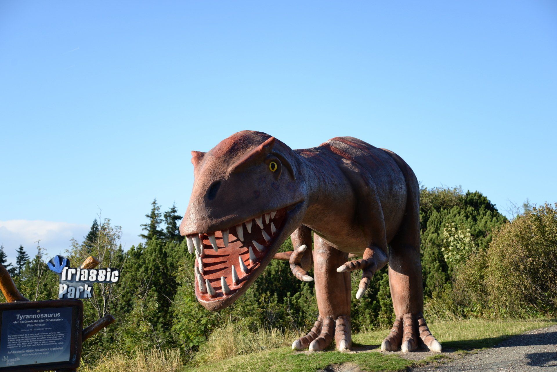 Eine große, braune T.-Rex-Statue steht im Freien vor blauem Himmel; in der Nähe befinden sich ein Schild und Büsche.