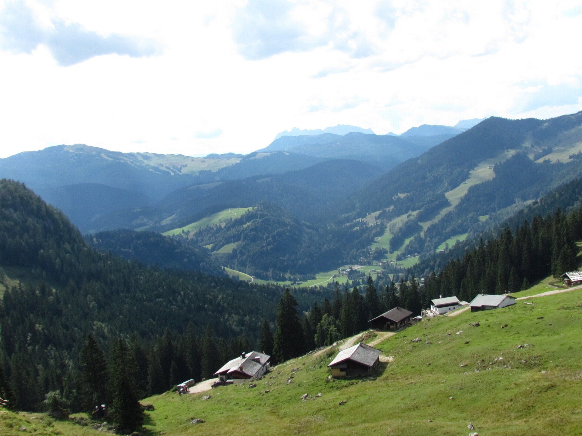 Bergtal mit bewaldeten Hügeln, Berghütten und einem grasbewachsenen Hang unter strahlend blauem Himmel