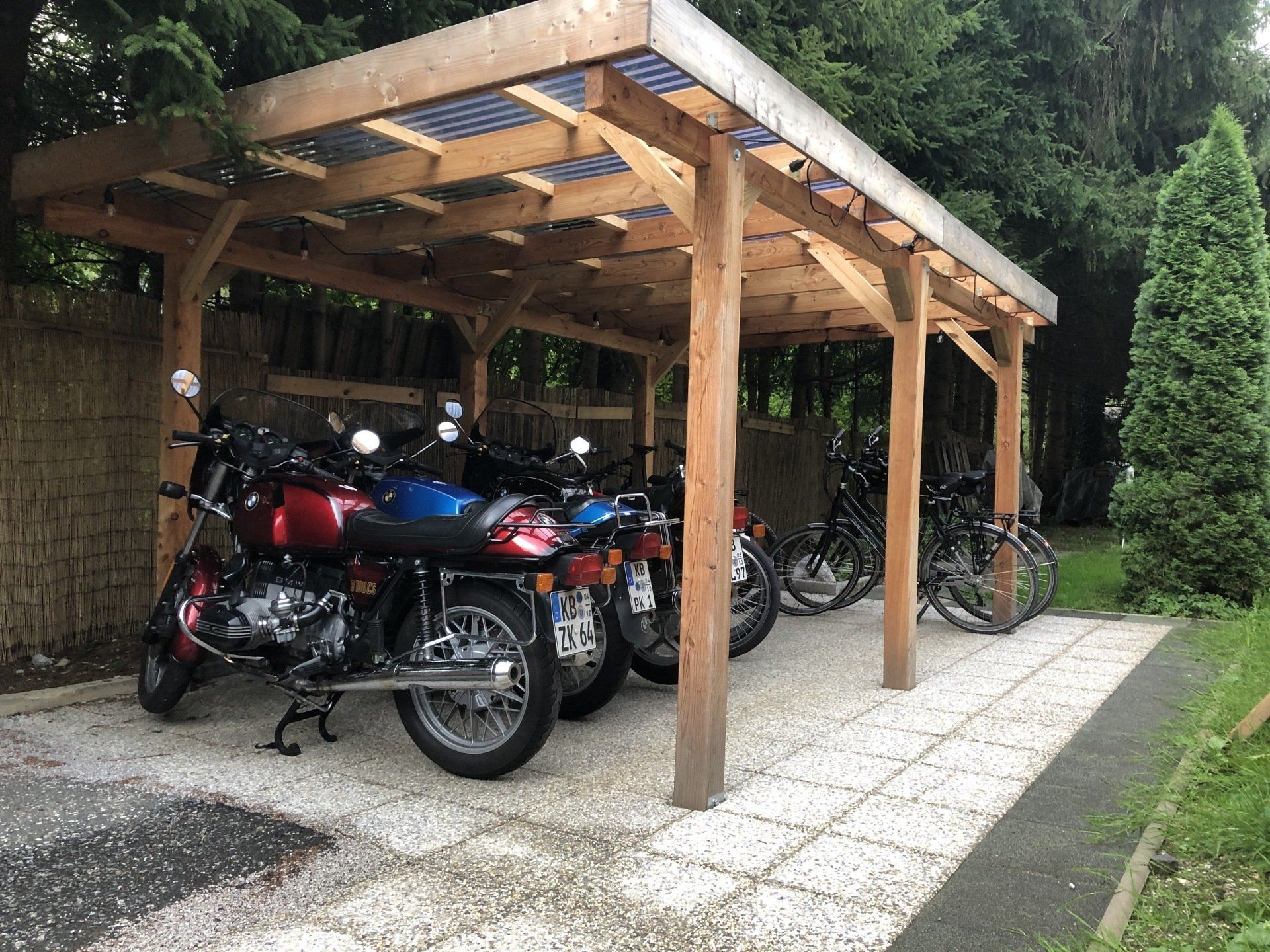 Überdachter Motorradparkplatz mit mehreren Motorrädern unter einem Holzunterstand neben einem Schotterweg