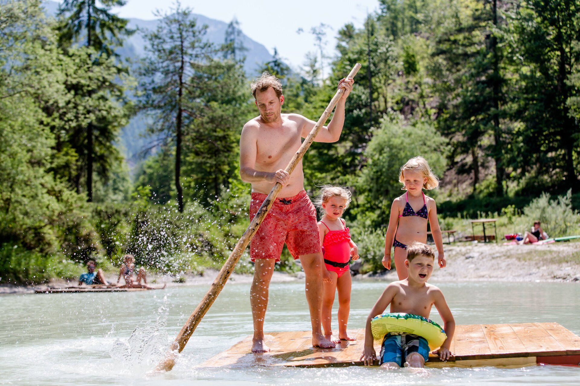 Menschen stehen auf einem Floß und rudern über einen Bergsee; Kinder sind in der Nähe.