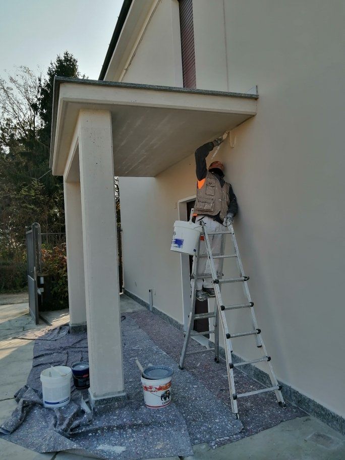 Persona su una scala che dipinge il soffitto del portico di un edificio; secchi di vernice e teloni a terra.