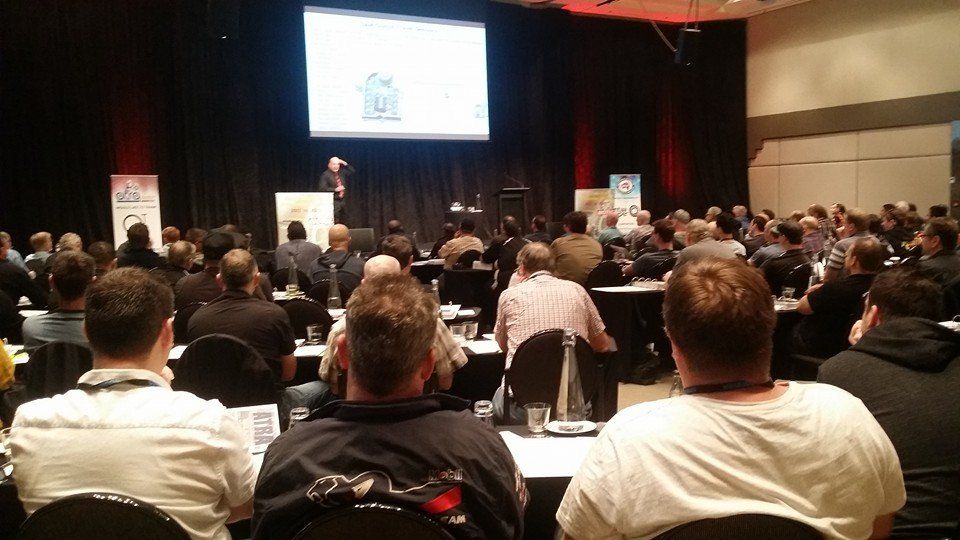Audience at a conference listens to a presenter on stage, a screen shows a house icon, indoor setting.