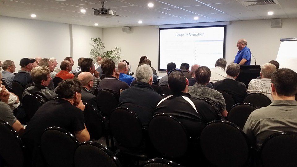 A man presenting to a seated audience in a conference room with a projector screen.