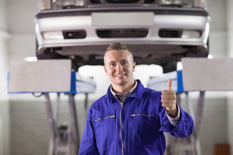 Mechanic in blue jumpsuit gives thumbs up under a car raised on a lift in a garage.