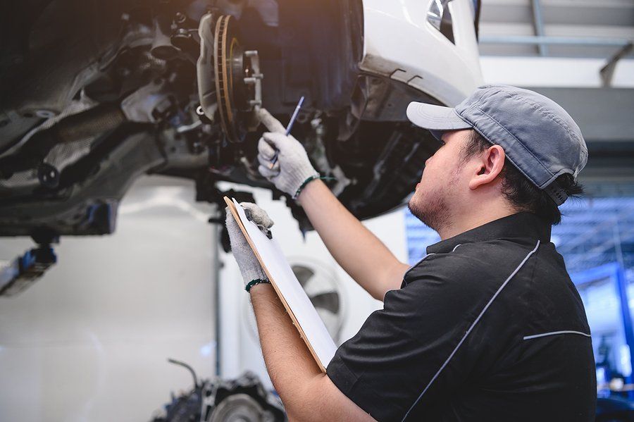 Mechanic inspecting car brakes, holding clipboard and pen. Wearing hat, gloves, and work attire in a garage.