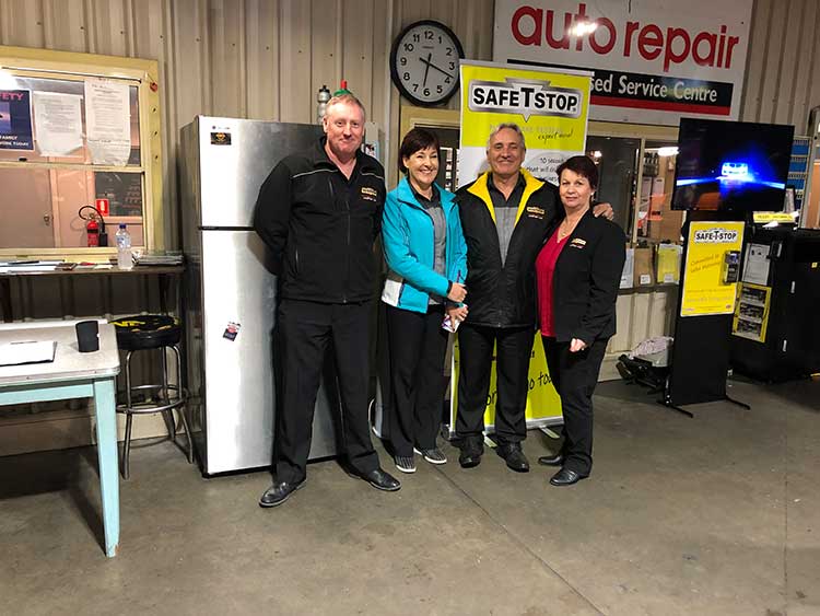 Four people pose in an auto repair shop. They stand near a refrigerator and a Safestop banner.