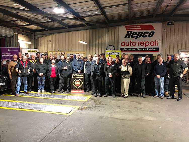 Large group of people posing in a Repco auto repair shop; banner and service bay visible.