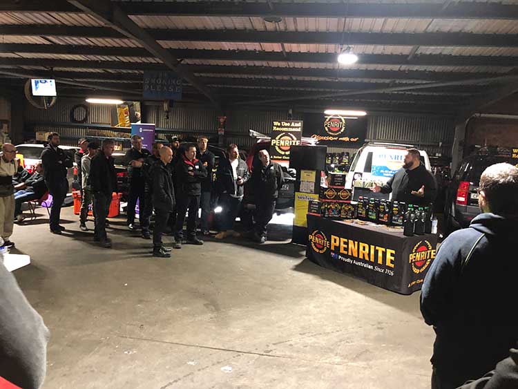 Group of people gathered at an event in a garage, a man speaking near a Penrite product display.
