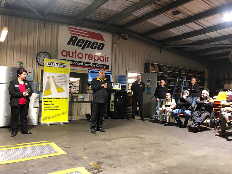 People at a Repco auto repair shop. A man speaks, others listen. Interior setting with tools and signage.