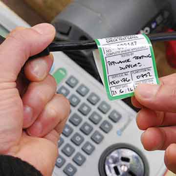 Hands holding an electrical cable with a tag, likely for appliance testing. A testing machine is visible in the background.