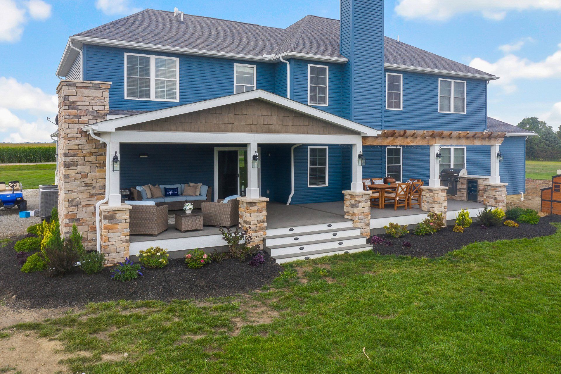 Blue two-story house with stone columns, covered porch, and green lawn under a cloudy sky