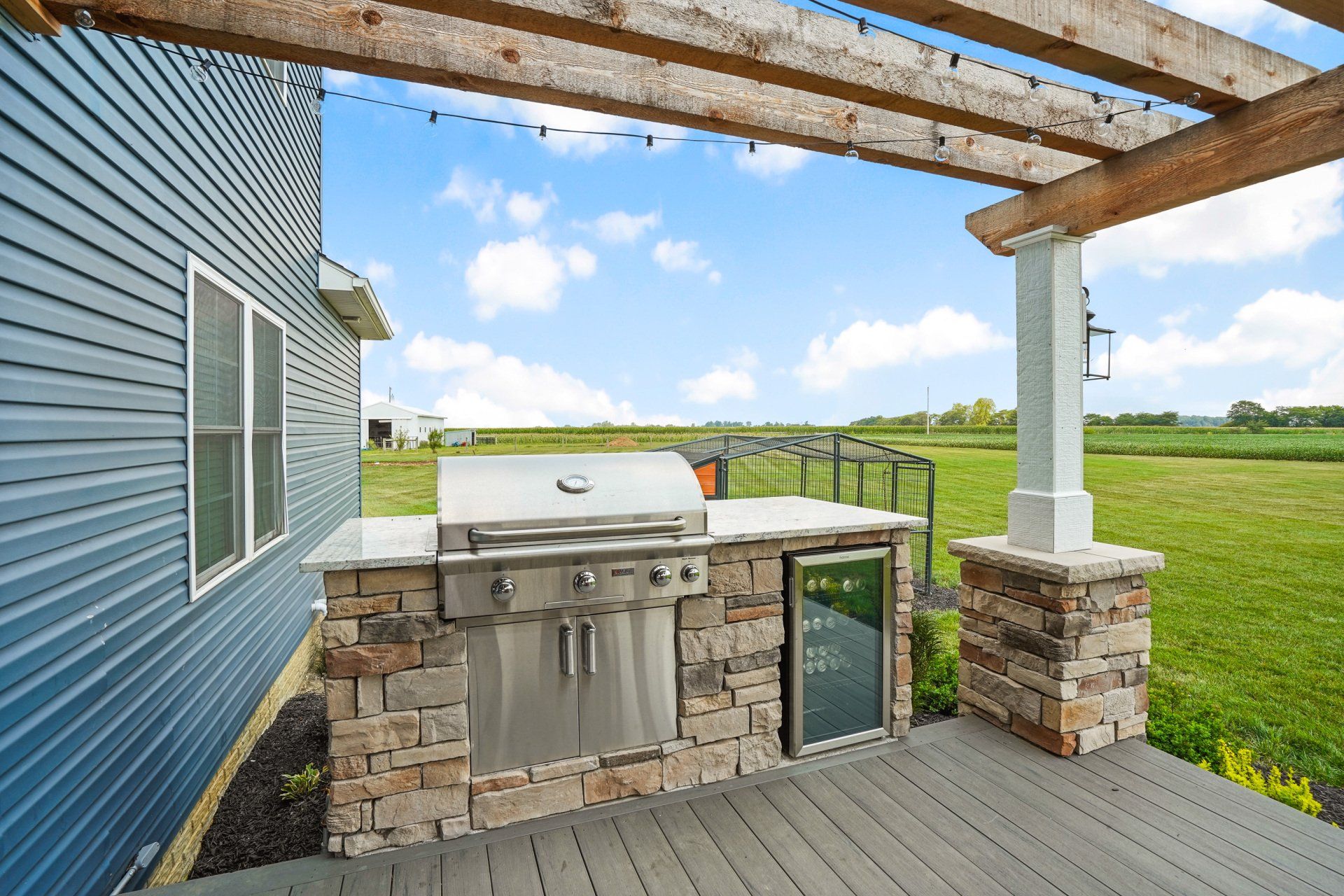 Outdoor stone grill on a wooden deck beside a blue house, overlooking a grassy field.
