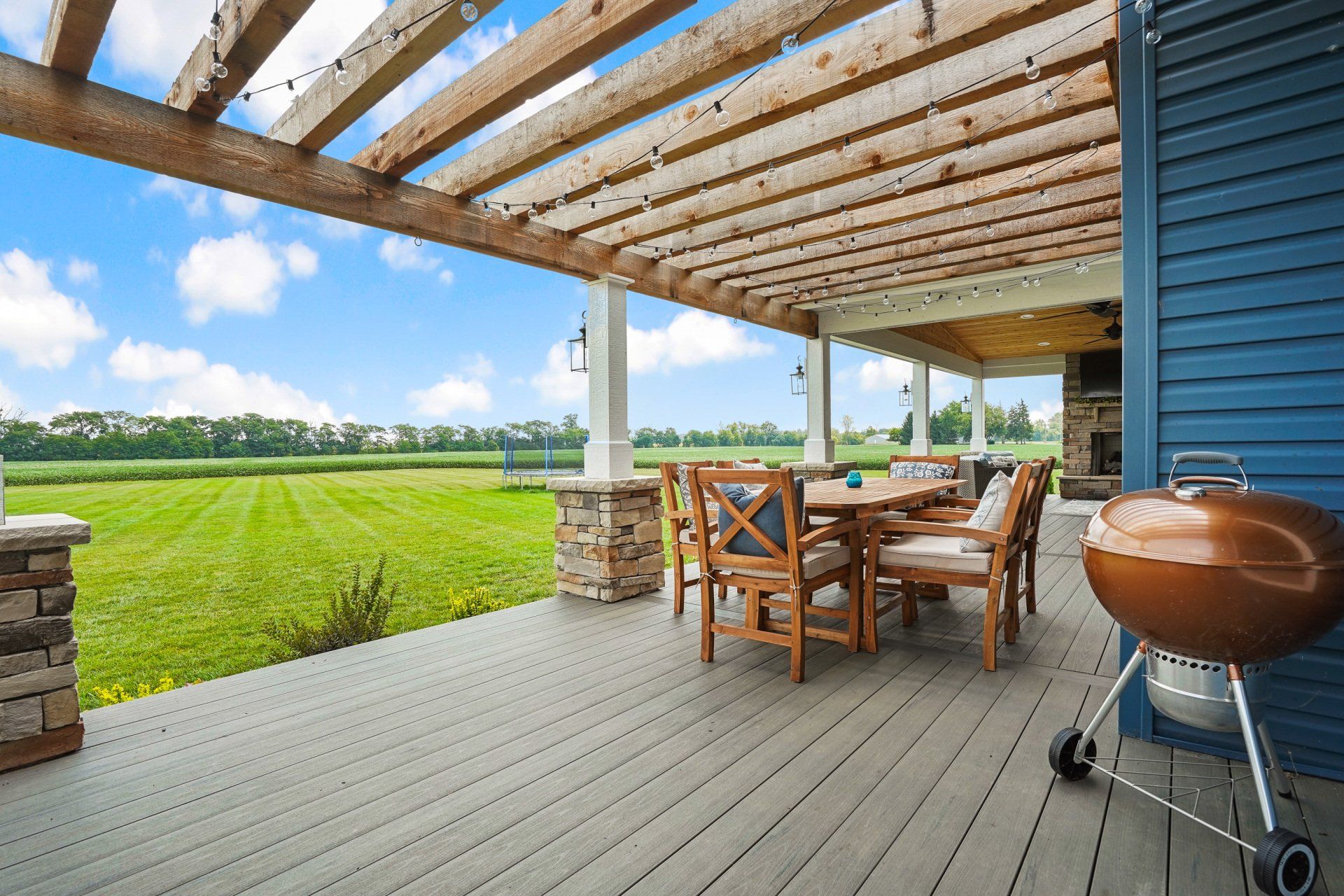 Covered patio with wooden dining set and grill overlooking a green lawn and open field