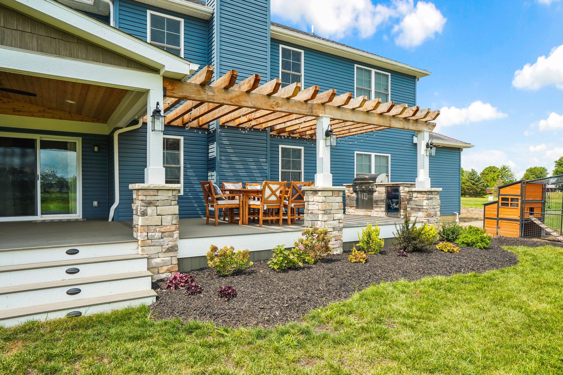 Backyard patio with pergola, outdoor dining set, and stone columns beside a blue house