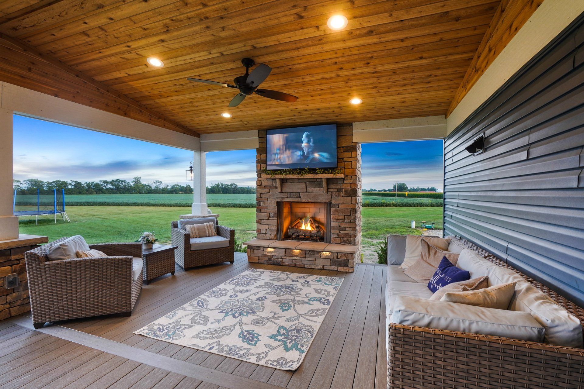 Covered patio with wicker seating, fireplace, TV, and view of green fields at sunset