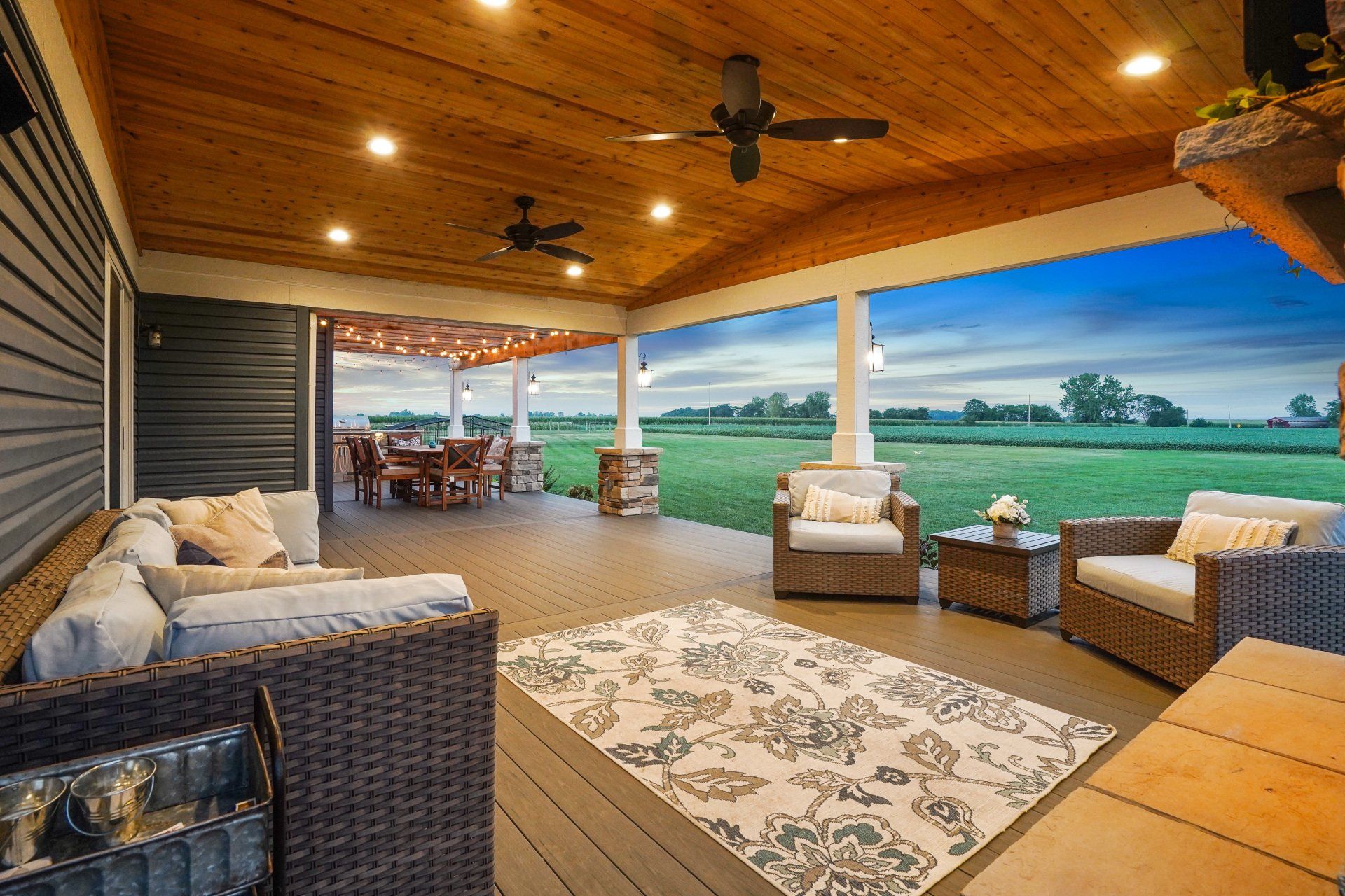 Covered wooden patio with wicker seating, ceiling fans, and a view of open green fields at sunset