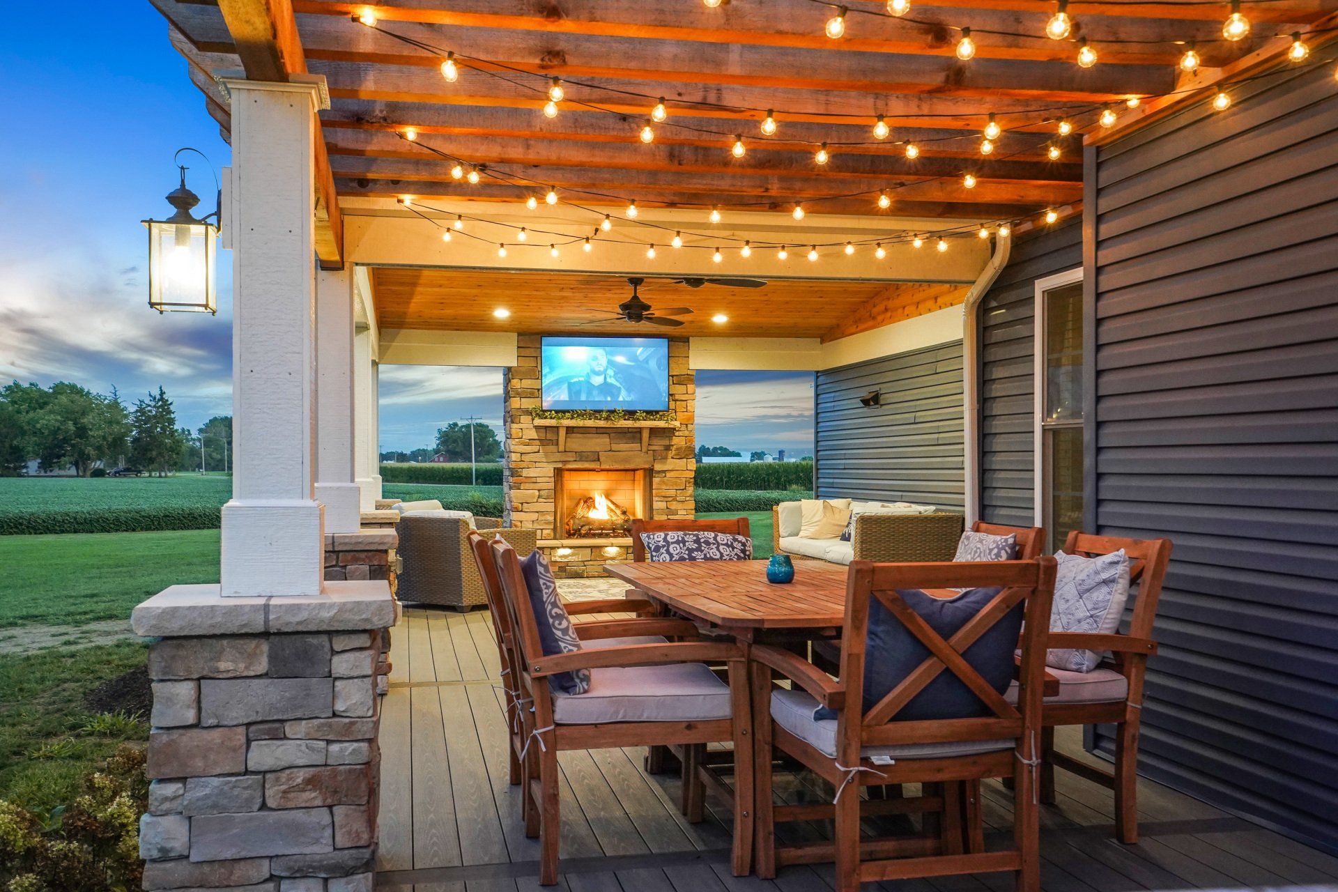 Covered wooden patio with string lights, outdoor dining table, and sunset view over a grassy landscape.