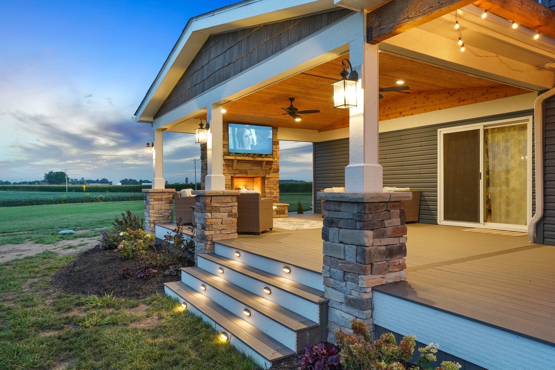 Covered farmhouse porch at dusk with stone columns, warm lights, and open countryside views