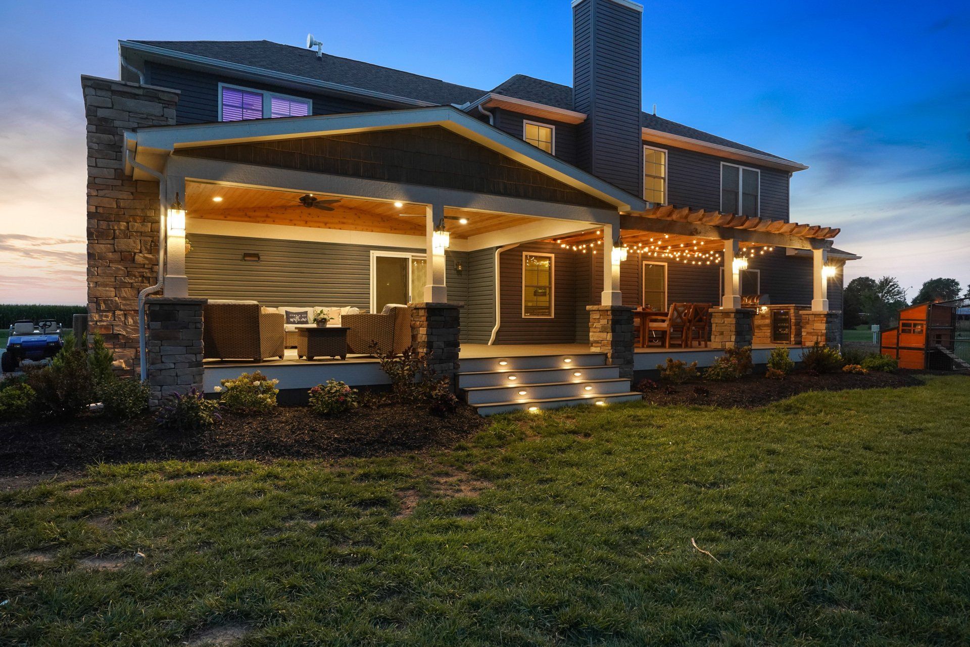 Modern two-story house with warm porch lights at dusk, featuring stone columns and a covered patio