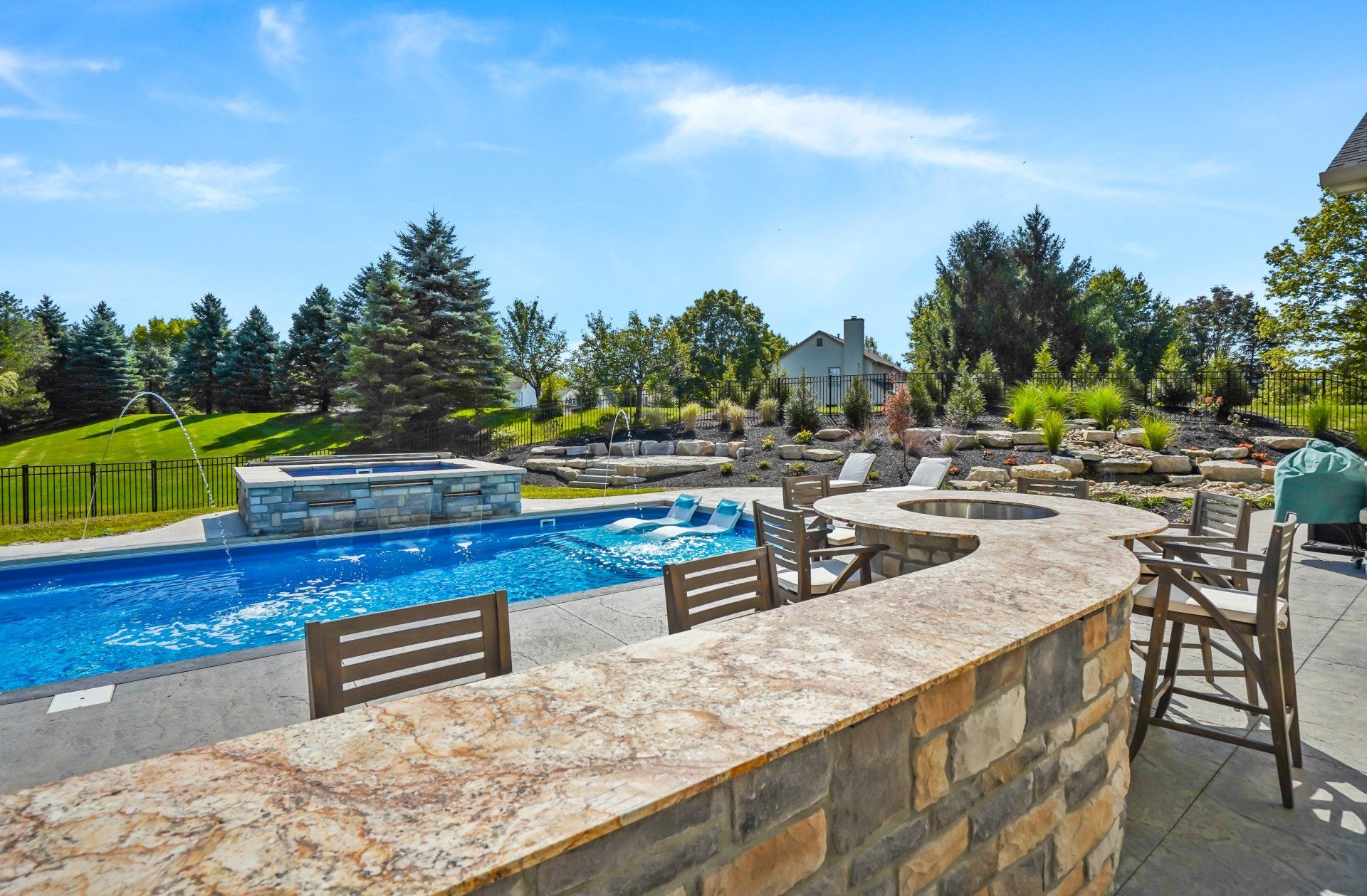 Stone poolside patio with blue swimming pool, lounge chairs, and landscaped garden under a sunny sky